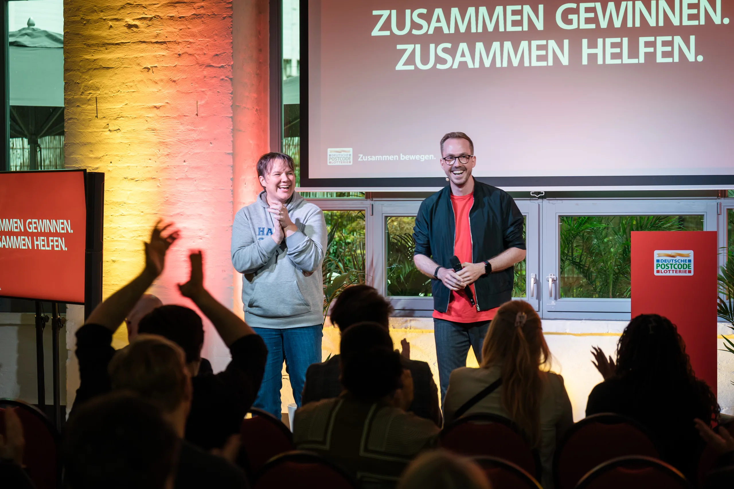 Applaus im Publikum beim Partnertreffen der Deutschen Postcode Lotterie 2025 in Berlin, Felix Uhlig vor der Leinwand mit dem Motto Zusammen gewinnen, zusammen helfen