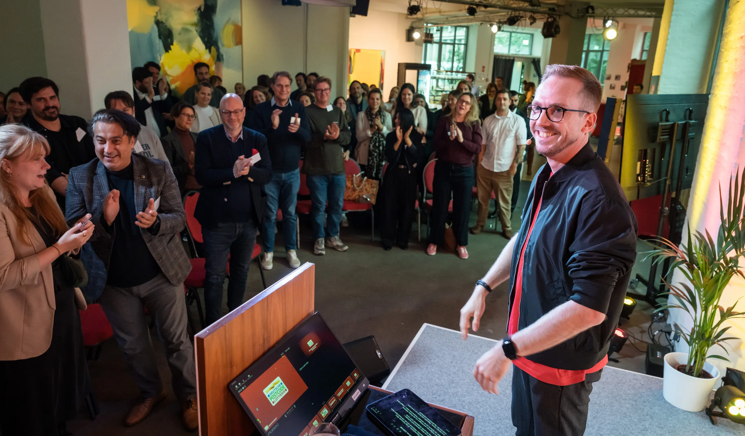 Felix Uhlig moderiert vor stehendem Publikum beim Partnertreffen der Deutschen Postcode Lotterie 2025