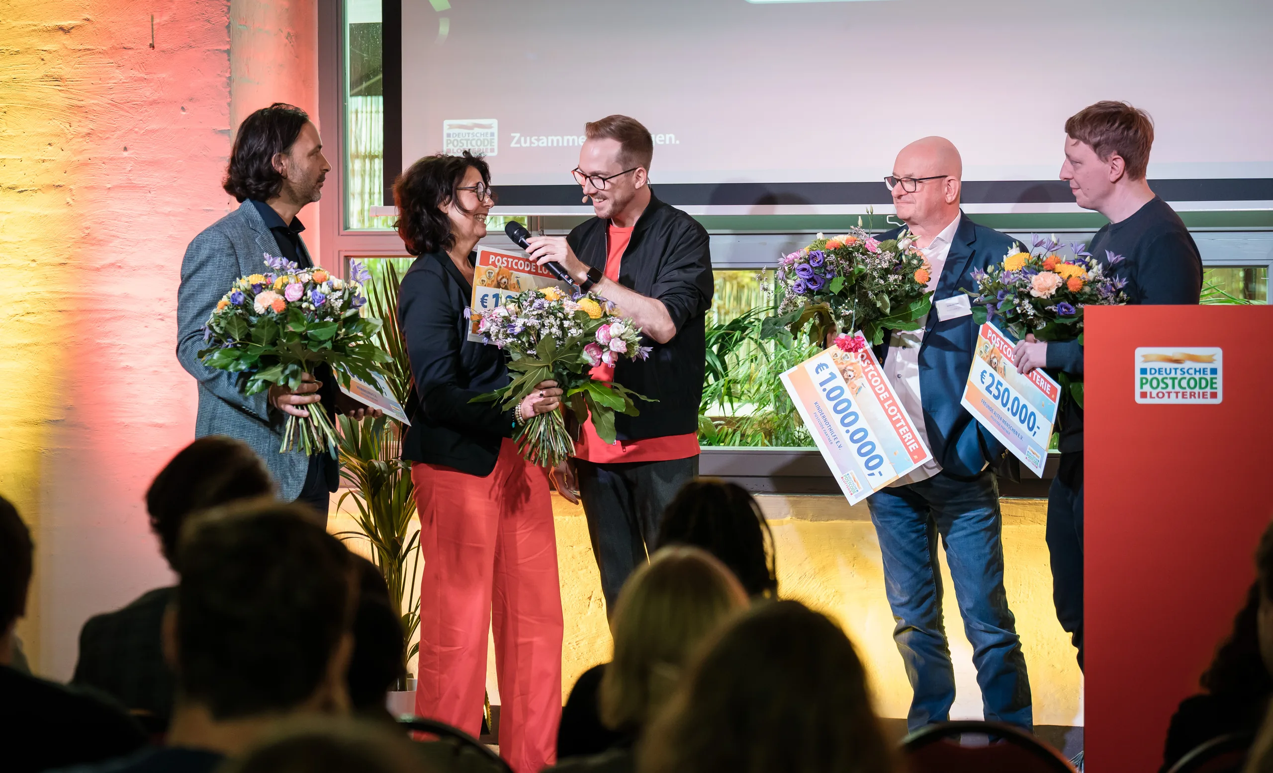 Felix Uhlig moderiert eine Scheckübergabe mit Blumen beim Partnertreffen der Deutschen Postcode Lotterie 2025